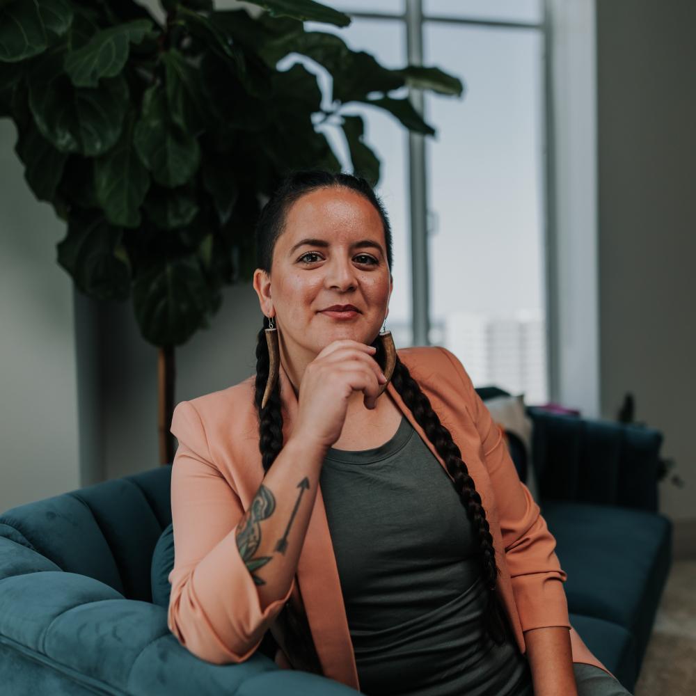 	This is a picture of Emi Aguilar of the Indigenous Cultures Institute. Emi is seated on a blue couch in front of a tree and window in an interior setting.