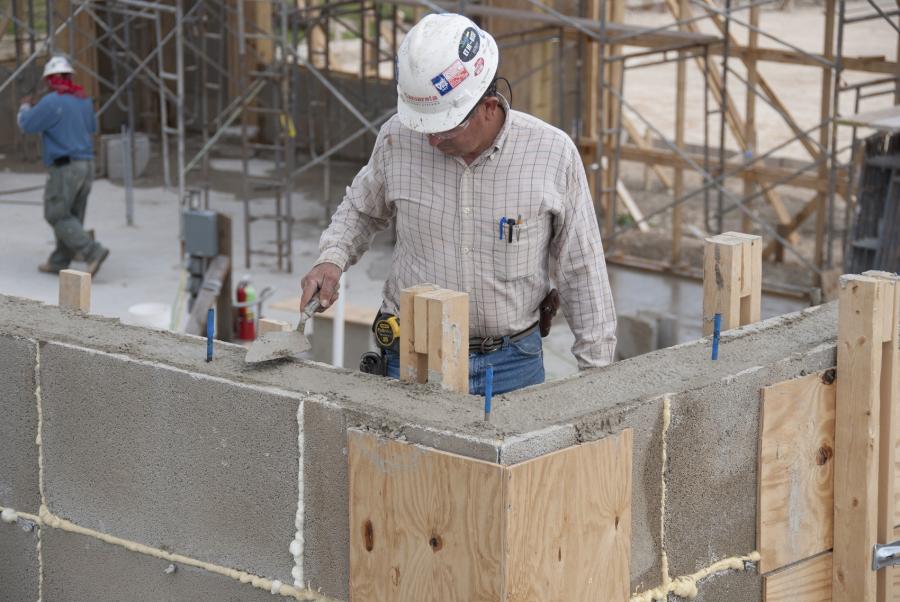 Construction worker wearing a white hard hat uses trowel to smooth mortar on the top of a concrete block wall. 