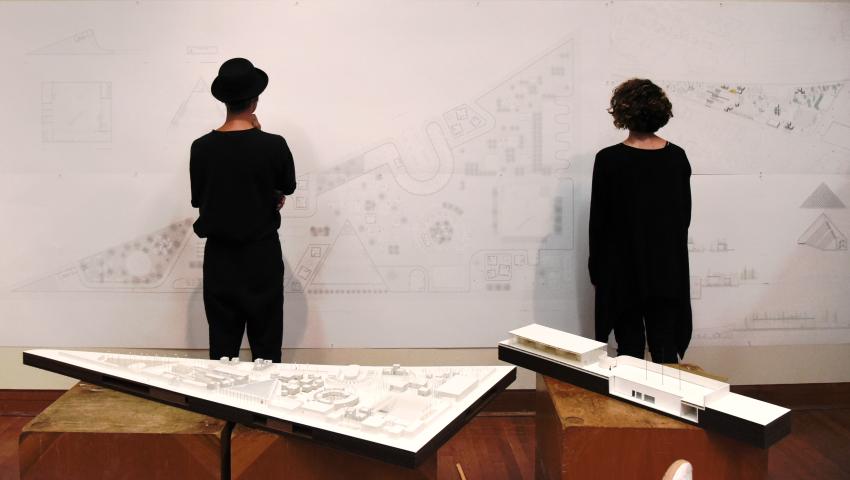Two students in black studying a pin-up with two large architecture models in the foreground