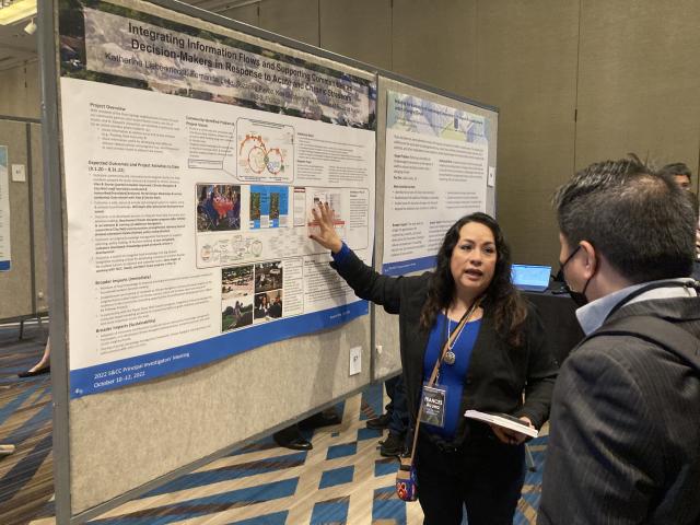 A woman stands in front of a poster on display at a National Science Foundation meeting. Her right hand is held up in front of the poster as she faces someone listening to her speak.