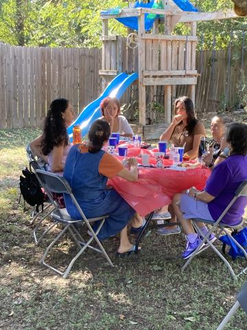 A group of six women sitting around a table with a pink tablecloth. They're outside in a backyard in front of a child's playhouse and slide.