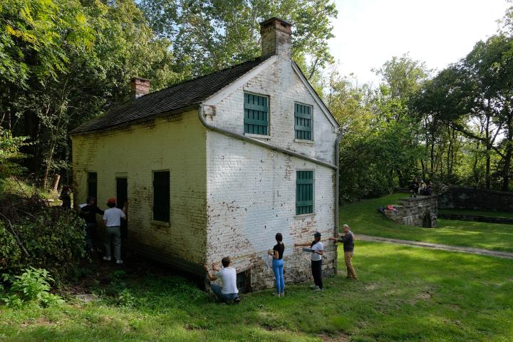 Four students seen measuring the side of a historic structure with a few others in the shade in the back of the building.