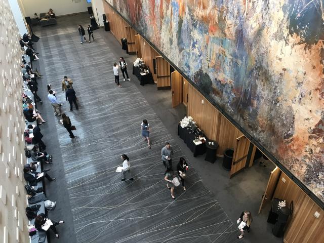 An overhead shot looking down at the lobby to the Zlotnik Family Ballroom. Students are seen sitting along a wall on the left side while others mingle and walk about in the lobby. On the right is a large-scale mural by Jose Parla.