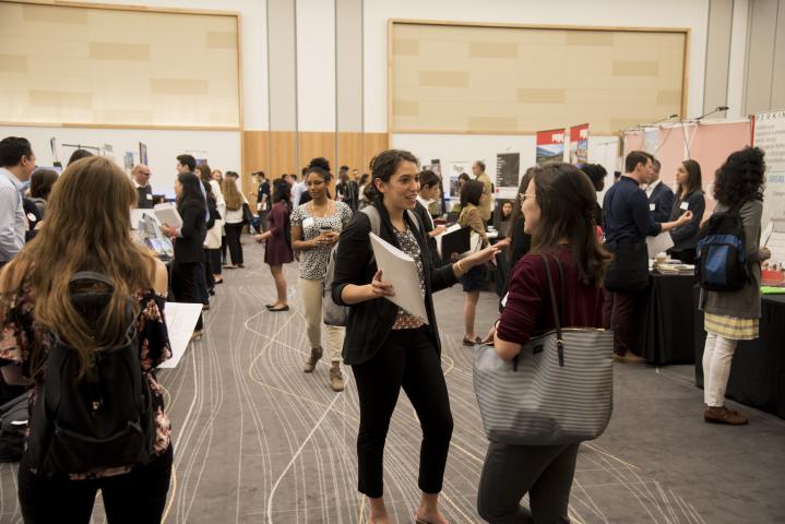 A student talks animatedly with another student in an aisle at the School of Architecture's Career Fair. Around them, many other students speak with employer representatives at boots on either side of the aisle. 