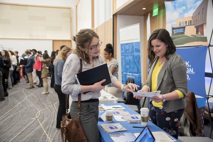 A female student holding a notebook holds out a resume as she talks to an employer set up at a booth at the School of Architecture's Career Fair. A group of other students are seen in the background.