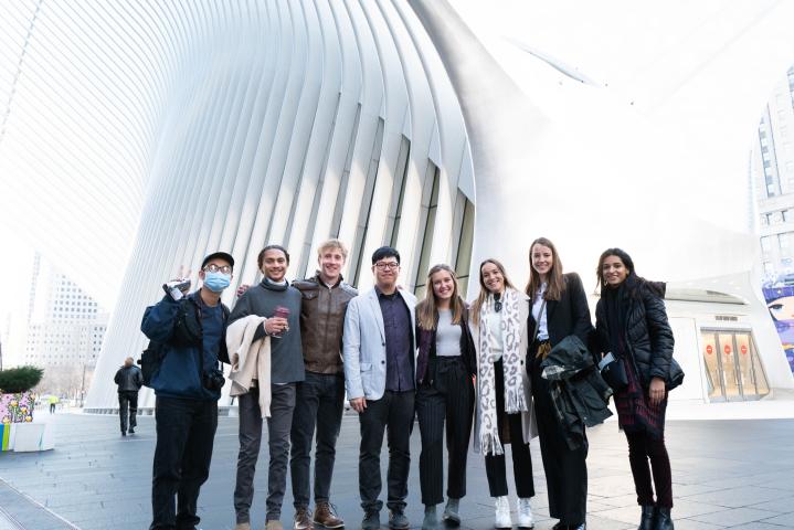 Eight students dressed up professionally smile at the camera in a group photo in front of an architectural building in New York City. One student is holding up a peace sign while wearing a mask.
